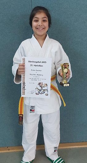 Ein lächelndes Kind in einem Weiß-Gelb-Judoanzug hält einen Pokal und eine Urkunde in den Händen. Die Urkunde bescheinigt den Gewinn des Bärchenpokals 2025 für den Judo-Club Hünfeld. Der Hintergrund ist blau und wirkt einladend.