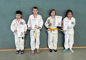 Vier Kinder in Judogewändern stehen nebeneinander und halten Urkunden in der Hand. Sie lächeln stolz. Einer der Jungen hält außerdem einen kleinen Pokal. Alle Kinder haben verschiedene Gürtel, die ihre Fortschritte im Judo zeigen. Der Hintergrund ist ein grauer Wandbereich.