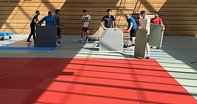 Eine Gruppe von sechs Personen trägt große Matten in einer Sporthalle. Die Matten bestehen aus verschiedenen Farben und sind teilweise auf einem roten Boden ausgelegt. Lichtstrahlen fallen durch Fenster und erzeugen Schatten auf dem Boden.
