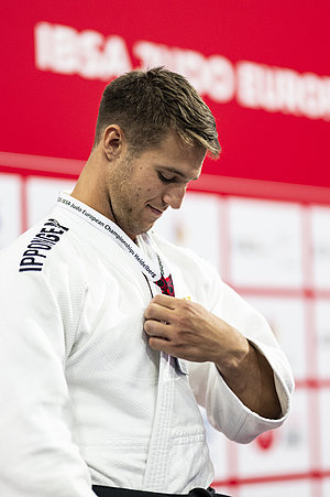 Ein junger Judoka in einem weißen Gi schaut auf seine Brust, während er eine Medaille oder Auszeichnung mit einem Lanyard befestigt. Der Hintergrund ist rot, mit unleserlichen Zeichen. Der Fokus liegt auf dem Athleten und dem Anbringen der Medaille.
