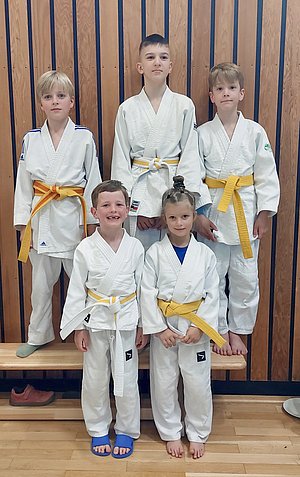 Fünf Kinder in Judogis stehen auf einer Bank. Zwei Kinder tragen gelbe Gürtel, eines hat einen weißen Gürtel. Die Kinder lachen und posieren freundlich. Im Hintergrund sind Holzpaneele zu sehen.