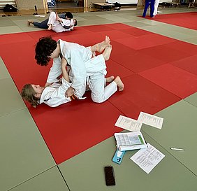Zwei Judoka üben auf einer roten Matte. Ein Junge befindet sich in oberer Position und greift die Umgebung der anderen Person, die auf dem Rücken liegt und lächelt. Im Hintergrund sind weitere Judokas in verschiedenen Positionen zu sehen.