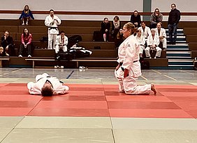Auf der Matte trainieren zwei Judokas. Eine Person kniet, während die andere auf dem Rücken liegt. Die Umgebung scheint eine Sporthalle zu sein, mit sitzenden Zuschauern im Hintergrund. Der Fokus liegt auf der Interaktion zwischen den Kämpfern.