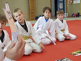 Vier Kinder in weißen Judoanzügen sitzen auf einer roten Matte und schauen aufmerksam in die Richtung eines ausgestreckten Papiers mit Augen. Sie scheinen an einer Aktivität oder Erzählung teilzunehmen. Im Hintergrund sind einige weitere Gegenstände unscharf sichtbar.