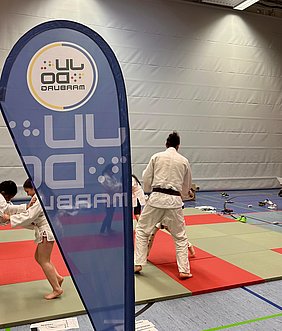 Auf dem Bild sind Kinder und Erwachsene zu sehen, die Judo praktizieren. Sie tragen weiße Judokimonos und üben auf einer Matte mit roten und grünen Bereichen. Im Hintergrund ist ein Banner mit dem Logo von "Marrburg" zu erkennen.