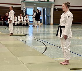 Eine junge Frau in einem weißen Judo-Anzug mit schwarzem Gürtel steht auf einer Judomatte. Im Hintergrund sind andere Judoka und Zuschauer zu sehen. Die Szene findet in einer Sporthalle statt, wo ein Judo-Training oder Wettkampf stattgefunden hat.