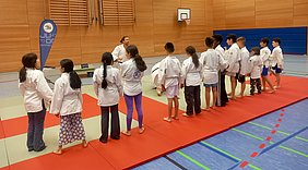 Eine Gruppe von Kindern in weißen Judo-Anzügen trennt sich in einer Sporthalle. Ein Trainer demonstriert eine Technik. Die Kinder beobachten aufmerksam, während einige auf roten Matten stehen. Es herrscht eine konzentrierte und freundliche Atmosphäre während des Trainings.