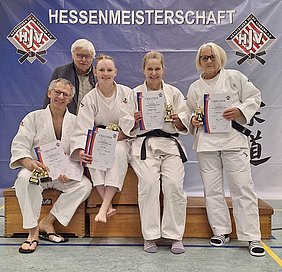 Fünf Personen in Judokleidung stehen zusammen auf einer Bühne, jede mit einer Urkunde und einer kleinen Trophäe. Sie zeigen stolz ihre Auszeichnungen von der Hessenmeisterschaft. Der Hintergrund ist mit einem Banner beschriftet, auf dem „Hessenmeisterschaft“ steht.