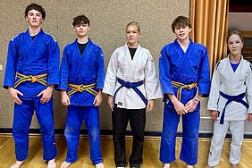 Vier Jugendliche stehen in Sportkleidung für Judo in einer Halle. Zwei tragen blaue Judogis mit farbigen Gürteln, während ein Mädchen einen weißen Judogi trägt und ein Junge schwarze Hosen. Die Gruppe posiert auf einem Holzboden vor einer neutralen Wand.