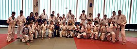 Eine Gruppe von Kindern und Erwachsenen in Judogis posiert fröhlich in einem Trainingsraum. Einige haben gelbe und blaue Gürtel. Die Gruppe zeigt verschiedene gestenreiche Posen. Im Vordergrund sitzt ein Teilnehmender mit einem Löwenkostüm.