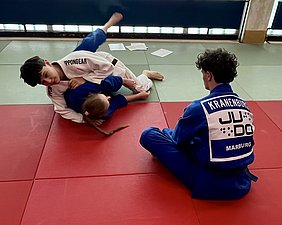 Drei Judokas üben auf einer Matte. Zwei kämpfen miteinander, während der dritte Zuschauer sitzt. Der Kämpfer in Weiß hat eine Technik angewendet, die andere Person in Blau liegt teilweise darunter. Die Matte ist rot und grün, und der Raum hat Tageslicht von Fenstern.
