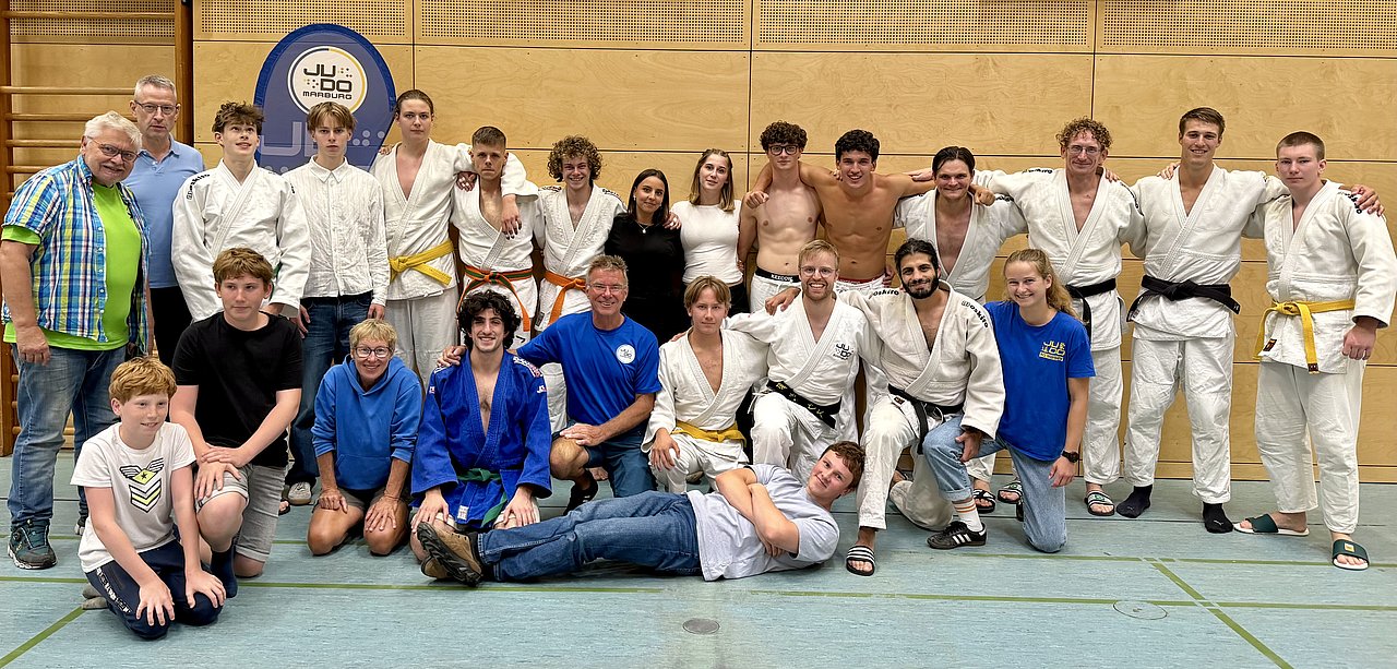 Eine Gruppe von Judokas posiert für ein Gruppenfoto in einem Sportzentrum. Sie tragen unterschiedliche Judo-Anzüge, und einige zeigen ihre Gürtelstufen. Im Hintergrund ist ein Judo-Vereinslogo sichtbar. Die Atmosphäre ist freundlich und sportlich.