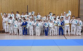 Eine Gruppe von Kindern in weißen Judo-Anzügen steht in einer Turnhalle, einige halten Medaillen und Urkunden in den Händen. Die Kinder lächeln und posieren stolz, während einige die Urkunden hochhalten. Im Hintergrund sind Holzpaneele und Matten zu sehen.