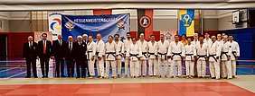 Gruppenfoto von Teilnehmern der Hessenmeisterschaft im Judo. Die Athleten tragen Judogis und Gürtel. Im Hintergrund sind Banner und Fahnen zu sehen. Alle Beteiligten stehen auf einem Judomatte, einige Personen haben den Blick zur Kamera gerichtet.