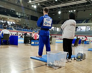 Ein junger Judoka in einem blauen Anzug steht aufmerksam auf einer Matte. Neben ihm steht ein Trainer in einer weißen Jacke mit dem Schriftzug "Deutschland". Im Hintergrund sind Zuschauer und Wettkämpfer zu sehen, die auf den Wettkampf im Judo fokussiert sind.