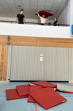 In einer Turnhalle stehen drei Jugendliche, zwei auf einem Balkon und einer davor. Ein Junge hebt eine rote Matte über seinen Kopf, während die anderen zuschauen. Auf dem Boden liegen mehrere rote Matten übereinander gestapelt.