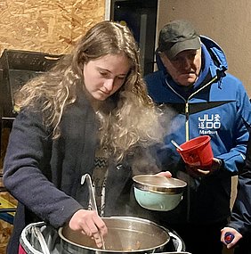 Eine junge Frau mit langen, lockigen Haaren rührt in einem großen Topf, aus dem Steam aufsteigt. Sie hält eine Schüssel in der Hand. Im Hintergrund steht ein älterer Mann in einer blauen Jacke und beobachtet sie aufmerksam.