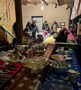Eine Gruppe von Menschen versammelt sich um Tische in einem geschützten Raum. Auf den Tischen sind verschiedene Speisen und Getränke wie Muffins, Kekse und Serviergeschirr sichtbar. Es herrscht eine gesellige Atmosphäre mit einer warmen Beleuchtung.