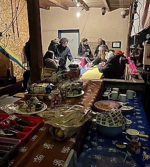 Eine Gruppe von Menschen versammelt sich um Tische in einem geschützten Raum. Auf den Tischen sind verschiedene Speisen und Getränke wie Muffins, Kekse und Serviergeschirr sichtbar. Es herrscht eine gesellige Atmosphäre mit einer warmen Beleuchtung.
