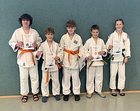 Fünf Kinder in Judokleidung stehen nebeneinander und halten Urkunden in der Hand. Sie zeigen stolz ihre Medaillen. Die Kinder sind lächelnd, einige tragen orange, grüne und weiße Gürtel. Der Hintergrund ist schlicht in einem grauen Farbton.