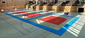 Eine Sporthalle mit mehreren Judomatten in verschiedenen Farben, hauptsächlich Rot und Blau, die auf dem Boden angeordnet sind. Lichtstrahlen fallen durch Fenster und erzeugen ein Muster auf den Matten. Stühle und eine kleine Tischanordnung sind am Rand sichtbar.