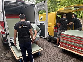 Vier Männer arbeiten im Freien, um große Platten in einen weißen Transporter und einen gelben Lieferwagen zu verladen. Die Platten sind grün und rot gestreift. Alle Männer tragen T-Shirts mit dem Logo „UNZÜGE“.