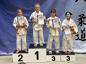 Vier Mädchen stehen auf dem Siegerpodest nach einem Judo-Wettkampf. Die Siegerin steht ganz links mit einer Urkunde, gefolgt von zwei weiteren Mädchen, die ebenfalls Urkunden und Medaillen halten. Das dritte Mädchen hält ein Stofftier. Der Hintergrund ist blau.