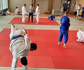 In einem Judotraining sind mehrere Personen auf rot-weißen Matten aktiv. Ein Judoka mit blauer Uniform führt einen Wurf aus, während andere Judokas in verschiedenen Aktivitäten beschäftigt sind. Die Umgebung scheint ein Sportzentrum zu sein.