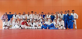 Gruppenfoto von Judoka in einem Dojo. Die Sportler tragen verschiedene Judoanzüge in Weiß, Blau und Schwarz. Sie stehen und sitzen zusammen auf einer roten Matte vor einer orangefarbenen Wand. Alle Teilnehmer zeigen lächelnde Gesichter und posieren fröhlich für das Bild.