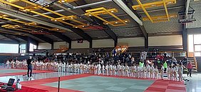 In einer Sporthalle stehen mehrere Kinder in weißen Judoanzügen in langen Reihen. Ein Trainer befindet sich vorne und spricht zu ihnen. Im Hintergrund sind Zuschauer auf Tribünen zu sehen. Der Boden ist mit einer matschenden Fläche für Judo bedeckt.