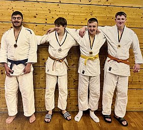 Vier Judoka stehen nebeneinander vor einer Holzwand. Sie tragen weiße Judoanzüge und Medaillen um ihren Hals. Der erste hat einen blauen Gürtel, der zweite einen grünen, der dritte einen gelben und der vierte einen orangefarbenen Gürtel. Sie lächeln und haben die Arme um die Schultern des Nachbarn gelegt.