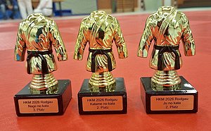 Drei goldene Trophäen in Form von Judokas, die auf schwarzen Sockeln stehen. Die Trophäen sind mit den Plätzen ausgezeichnet: links "3. Platz, Nage no kata", in der Mitte "2. Platz, Katame no kata", rechts "2. Platz, Ju no kata". Hintergrund in Rot.